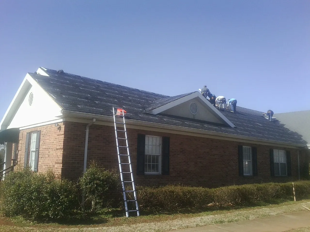 Crew tearing shingles off a brick ranch roof with a ladder set — Metal Roof Repair work by our crew in The Villages