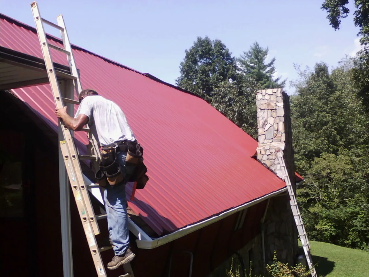 Roofer on a ladder beside a red metal roof and stone chimney — Soffit & Fascia Repair by Roofing Brothers in The Villages